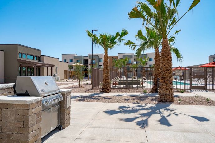 Mesa Vista Affordable Apartments with Outdoor grill near palm trees with a gated pool and modern apartment buildings in the background under a clear sky.