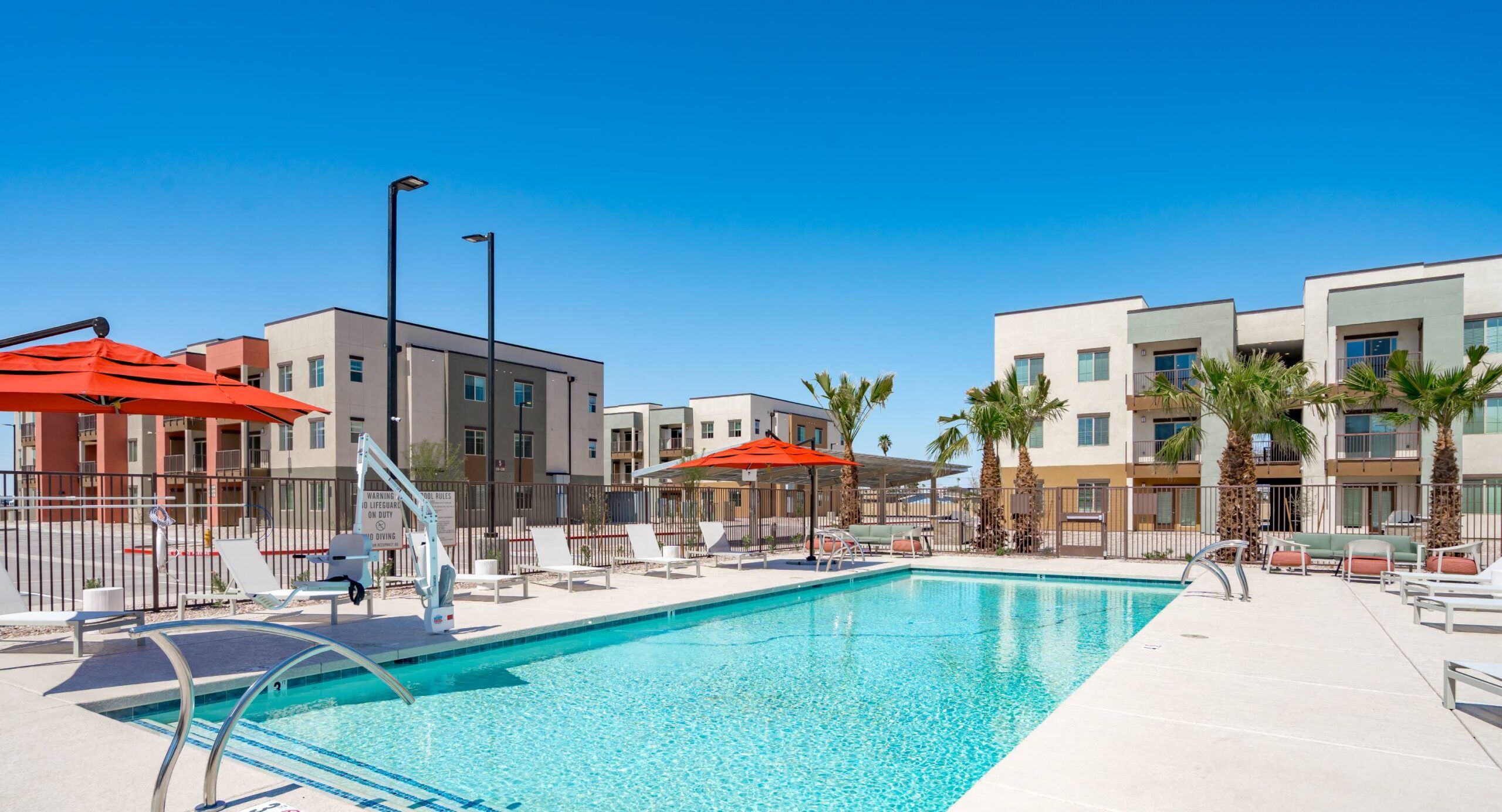 Outdoor swimming pool with lounge chairs, red umbrellas, and modern apartment buildings in the background.