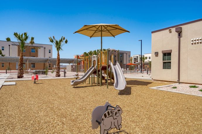 Mesa Vista Affordable Apartments with Playground with slides, shade umbrella, toy dinosaur, and palm trees, surrounded by buildings on a sunny day.