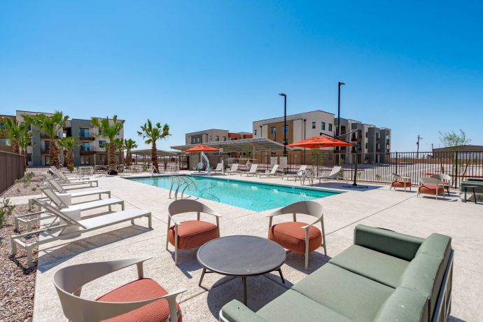 Mesa Vista Affordable Apartments with Outdoor pool area with lounge chairs, tables, and umbrellas, surrounded by modern apartment buildings on a sunny day.